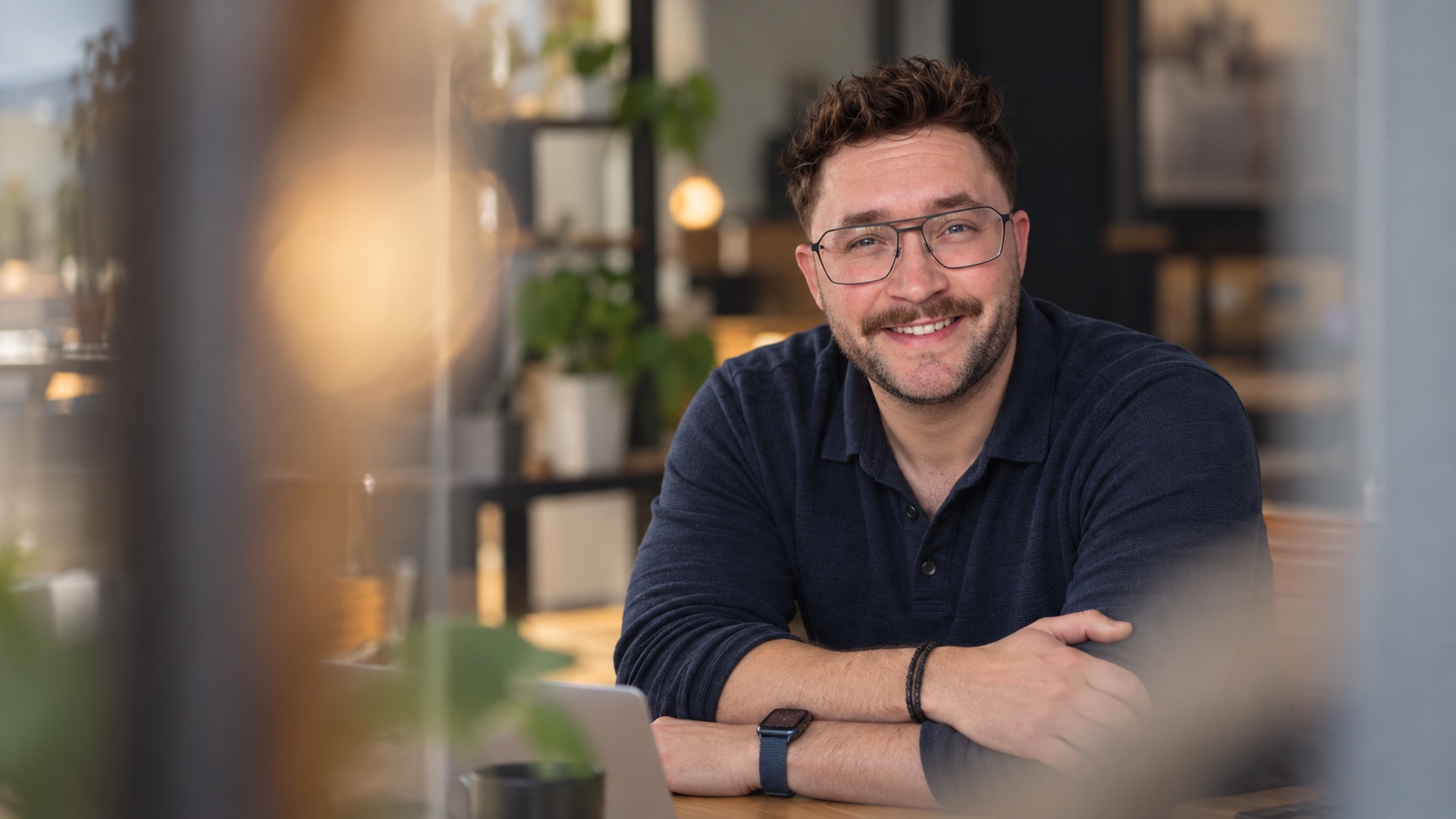 Ein Porträt-Foto von Christoph Hampe, der lächelnd an einem Holztisch in einem modernen Co-Working-Space sitzt. Er trägt eine Brille, ein blaues Poloshirt und eine Smartwatch.